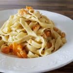 A white plate holds a serving of fettuccine pasta mixed with chunks of roasted butternut squash. The plate is placed on a wooden table, and the pasta is arranged in a small mound, showcasing the creamy texture of the pasta and the vibrant orange of the squash.