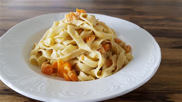 A white plate holds a serving of fettuccine pasta mixed with chunks of roasted butternut squash. The plate is placed on a wooden table, and the pasta is arranged in a small mound, showcasing the creamy texture of the pasta and the vibrant orange of the squash.