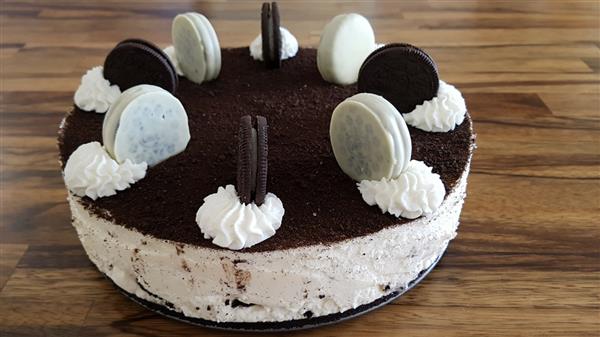 A round cookies and cream cheesecake sits on a wooden surface. It is topped with a layer of crushed cookies, dollops of whipped cream, and alternating whole chocolate and white chocolate sandwich cookies placed upright.