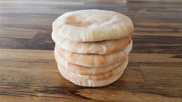 A stack of six round, golden-brown pita breads is placed on a wooden surface. The bread is slightly puffed and looks soft and fresh, with a few visible flour spots. The wood features a natural grain pattern and varying shades of brown.