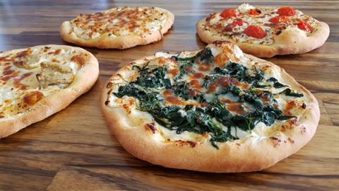 Four fresh, homemade pizzas on a wooden surface. The pizzas have various toppings: one with green spinach and melted cheese, another with cherry tomatoes and cheese, a third with slices of chicken, and a fourth with a simple cheese topping.