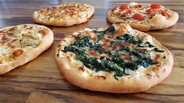 Four fresh, homemade pizzas on a wooden surface. The pizzas have various toppings: one with green spinach and melted cheese, another with cherry tomatoes and cheese, a third with slices of chicken, and a fourth with a simple cheese topping.