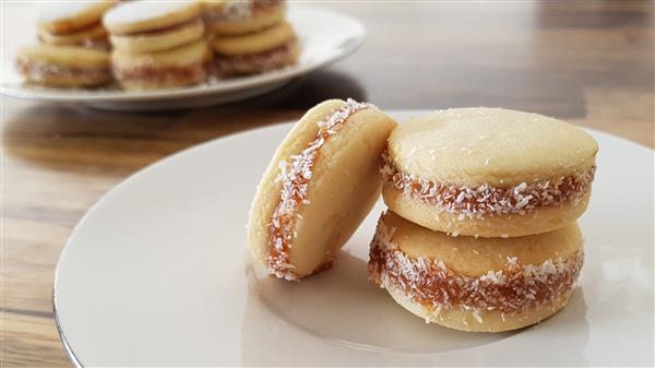 Two alfajores, sandwich cookies filled with dulce de leche and rolled in coconut, are stacked on a white plate. In the background, a plate with more alfajores is visible on a wooden table.