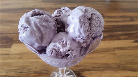 A close-up of a glass bowl filled with scoops of creamy, purple ice cream. The ice cream appears smooth and rich, set against a blurred wooden table in the background.