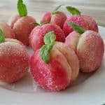 A plate of peach-shaped cookies coated in sugar, consisting of two halves joined together, with each cookie decorated with a small mint leaf on top. The cookies are pink and white, mimicking the appearance of real peaches. They are arranged on a white plate.