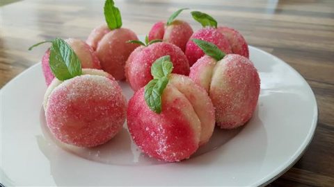 A plate of peach-shaped cookies coated in sugar, consisting of two halves joined together, with each cookie decorated with a small mint leaf on top. The cookies are pink and white, mimicking the appearance of real peaches. They are arranged on a white plate.