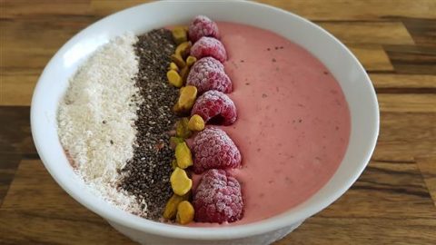 A bowl of pink smoothie topped with rows of white shredded coconut, dark chia seeds, frozen raspberries, and green pistachios. The bowl is set on a wooden surface.