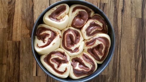 A round baking tray filled with unbaked, rolled dough pastries swirled with chocolate. The dough is light in color, and the chocolate forms distinct, dark swirls. The tray is placed on a wooden surface.