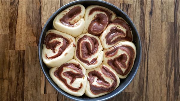 A round baking tray filled with unbaked, rolled dough pastries swirled with chocolate. The dough is light in color, and the chocolate forms distinct, dark swirls. The tray is placed on a wooden surface.