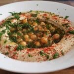 A white plate of creamy hummus garnished with whole chickpeas, fresh chopped parsley, a drizzle of olive oil, and sprinkled with red paprika. The dish is served on a wooden table.