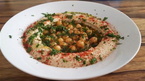 A white plate of creamy hummus garnished with whole chickpeas, fresh chopped parsley, a drizzle of olive oil, and sprinkled with red paprika. The dish is served on a wooden table.