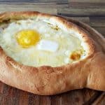 A freshly baked khachapuri on a wooden surface. The Georgian cheese-filled bread boat is topped with a sunny-side-up egg and a dollop of butter, giving it a rich and appetizing appearance. The crust is golden brown and surrounded by a warm, inviting ambiance.