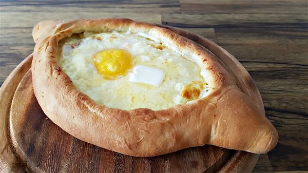 A freshly baked khachapuri on a wooden surface. The Georgian cheese-filled bread boat is topped with a sunny-side-up egg and a dollop of butter, giving it a rich and appetizing appearance. The crust is golden brown and surrounded by a warm, inviting ambiance.