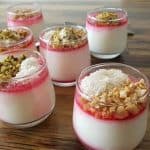 A wooden table holds six glass jars of layered yogurt parfaits. Each parfait has a bottom layer of white yogurt, a pink syrup layer in the middle, and a topping that includes various ingredients such as pistachios, shredded coconut, and chopped nuts. Silver spoons are placed around the jars.