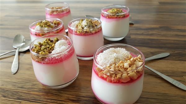 A wooden table holds six glass jars of layered yogurt parfaits. Each parfait has a bottom layer of white yogurt, a pink syrup layer in the middle, and a topping that includes various ingredients such as pistachios, shredded coconut, and chopped nuts. Silver spoons are placed around the jars.