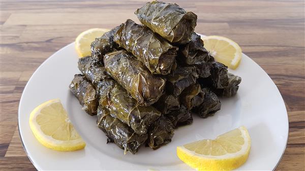 A plate of neatly stacked stuffed grape leaves (dolma), surrounded by six small lemon wedges. The dish is presented on a wooden surface.