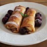 A white plate on a wooden table holds two rolled crepes, dusted with powdered sugar, and garnished with a small green herb. Dark red cherries are placed around the crepes, and syrup or sauce is drizzled on the plate.