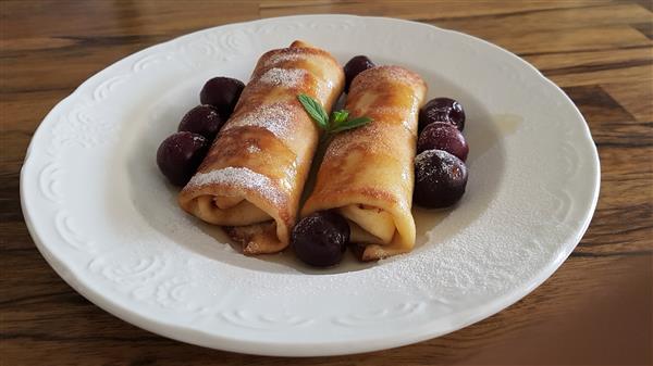 A white plate on a wooden table holds two rolled crepes, dusted with powdered sugar, and garnished with a small green herb. Dark red cherries are placed around the crepes, and syrup or sauce is drizzled on the plate.