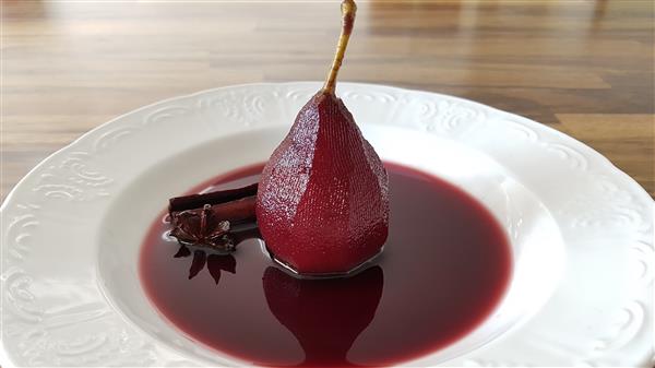 A poached pear sits upright in a white plate filled with red wine sauce. Next to the pear, there appears to be a piece of cinnamon stick and a star anise, adding to the dish's presentation. The background features a wooden surface.