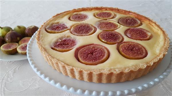 A fig tart sits on a decorative white plate. The tart has a golden-brown crust and is topped with fresh fig slices embedded in a creamy filling. In the background, whole figs are arranged on a separate white plate. The tablecloth is light-colored with a subtle pattern.