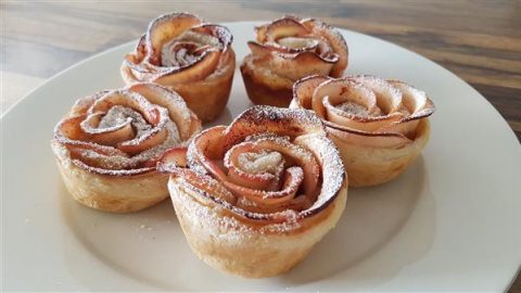 Five apple rose pastries sit on a white plate. Each pastry is intricately crafted to resemble a blooming rose, with thinly sliced apple pieces forming the petals. The pastries are lightly dusted with powdered sugar. The background features a wooden table.