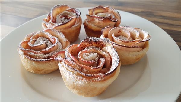 Five apple rose pastries sit on a white plate. Each pastry is intricately crafted to resemble a blooming rose, with thinly sliced apple pieces forming the petals. The pastries are lightly dusted with powdered sugar. The background features a wooden table.