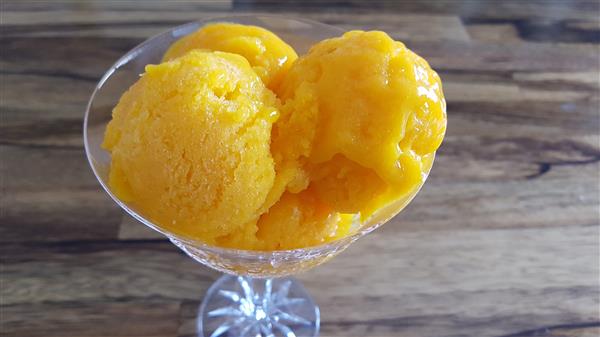 A glass dish containing three scoops of bright orange mango sorbet is set on a wooden surface.