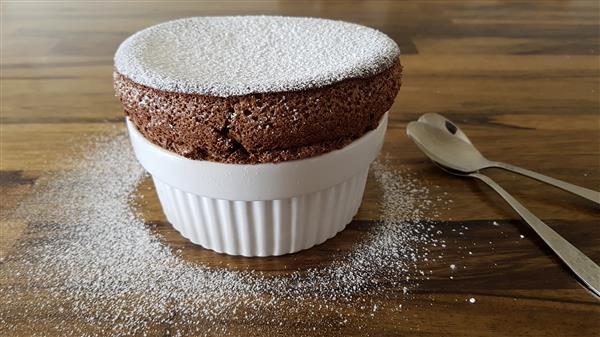 A chocolate soufflé dusted with powdered sugar sits in a white ramekin on a wooden surface. Two spoons rest beside the ramekin, and some powdered sugar is scattered around.