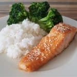 A white plate holds a meal of sesame-seed-topped salmon fillet, a serving of steamed white rice, and a portion of cooked broccoli florets. The plate is placed on a wooden surface.