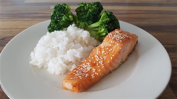 A white plate holds a meal of sesame-seed-topped salmon fillet, a serving of steamed white rice, and a portion of cooked broccoli florets. The plate is placed on a wooden surface.