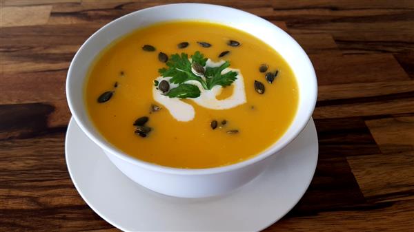 A white bowl filled with creamy pumpkin soup, garnished with a swirl of cream, parsley, and seeds. The bowl sits on a matching white saucer on a wooden table.