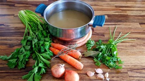 A steel pot filled with broth sits on a wooden countertop surrounded by fresh vegetables and herbs. Ingredients include celery stalks, carrots, parsley, onions, garlic cloves, and sprigs of thyme and bay leaves. The scene suggests preparing homemade soup or stock.