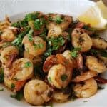 A white plate is filled with cooked shrimp garnished with fresh chopped herbs and served with a lemon wedge on the side. The shrimp appear to be seasoned with spices and herbs, and the dish is placed on a wooden surface.