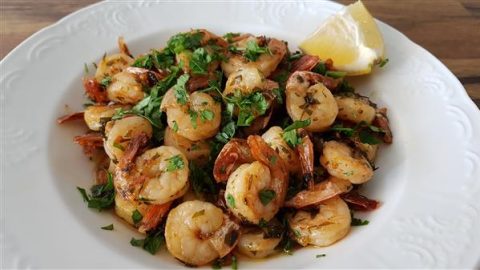 A white plate is filled with cooked shrimp garnished with fresh chopped herbs and served with a lemon wedge on the side. The shrimp appear to be seasoned with spices and herbs, and the dish is placed on a wooden surface.