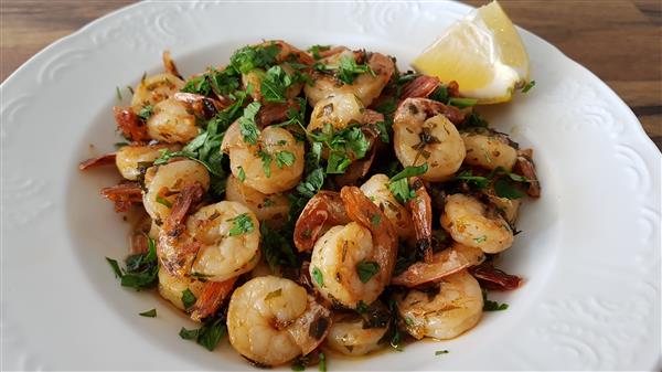 A white plate is filled with cooked shrimp garnished with fresh chopped herbs and served with a lemon wedge on the side. The shrimp appear to be seasoned with spices and herbs, and the dish is placed on a wooden surface.