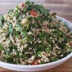 A bowl filled with a fresh quinoa salad sits on a wooden table. The salad is a colorful blend of cooked quinoa, chopped fresh parsley, diced tomatoes, and green herbs. The ingredients are finely mixed, creating a vibrant and healthy dish.