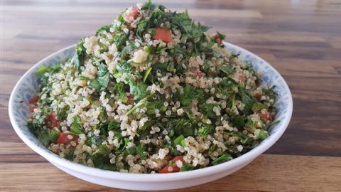 A bowl filled with a fresh quinoa salad sits on a wooden table. The salad is a colorful blend of cooked quinoa, chopped fresh parsley, diced tomatoes, and green herbs. The ingredients are finely mixed, creating a vibrant and healthy dish.