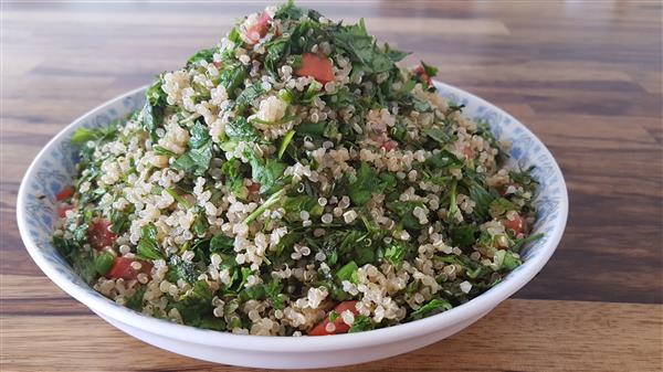 A bowl filled with a fresh quinoa salad sits on a wooden table. The salad is a colorful blend of cooked quinoa, chopped fresh parsley, diced tomatoes, and green herbs. The ingredients are finely mixed, creating a vibrant and healthy dish.