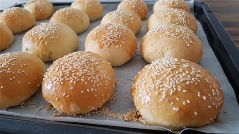 A baking tray filled with freshly baked, golden brown burger buns topped with white sesame seeds, resting on a sheet of parchment paper.