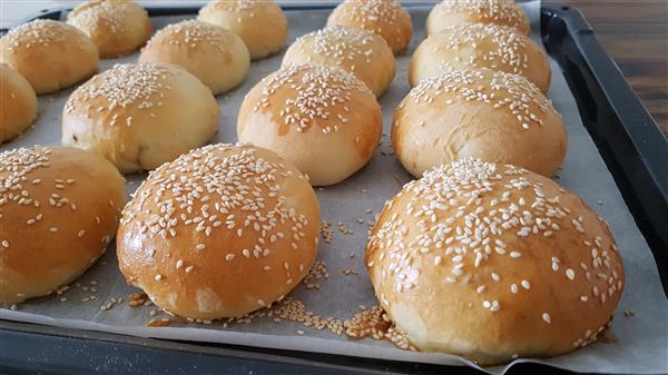 A baking tray filled with freshly baked, golden brown burger buns topped with white sesame seeds, resting on a sheet of parchment paper.