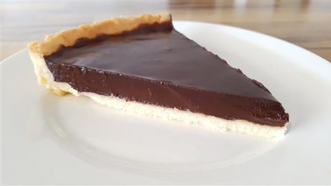 A slice of chocolate tart with a smooth, glossy surface and a light, flaky crust is placed on a white plate. The tart has a rich, thick chocolate filling. The background shows a wooden table.