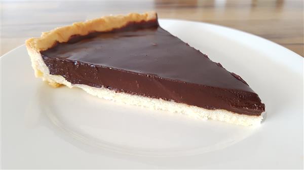 A slice of chocolate tart with a smooth, glossy surface and a light, flaky crust is placed on a white plate. The tart has a rich, thick chocolate filling. The background shows a wooden table.