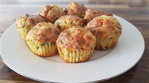 A plate of golden-brown savory muffins with visible herbs and cheese. The muffins are arranged in a circular pattern on a white plate, placed on a wooden table.