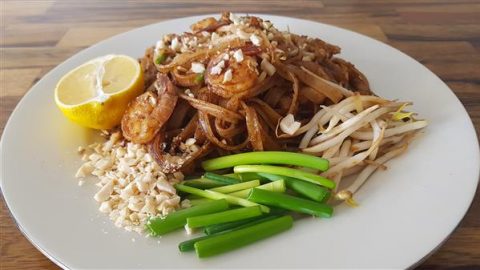 A plate of pad Thai garnished with chopped peanuts, a lemon wedge, bean sprouts, and green onions. The dish includes stir-fried noodles with shrimp and is placed on a white plate on a wooden table.