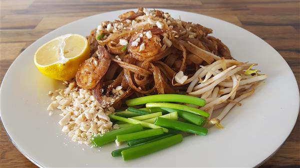 A plate of pad Thai garnished with chopped peanuts, a lemon wedge, bean sprouts, and green onions. The dish includes stir-fried noodles with shrimp and is placed on a white plate on a wooden table.