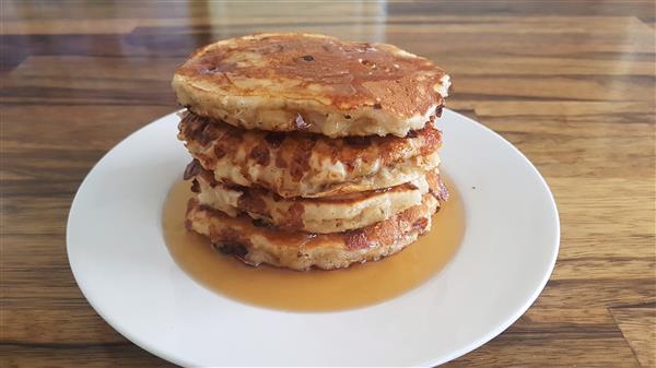 A stack of four golden-brown pancakes sits on a white plate, drizzled with syrup. The pancakes have a slightly crispy exterior and are thick and fluffy. The plate is placed on a wooden table, and the syrup pools around the base of the stack.