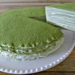 A matcha green tea crepe cake is placed on a white plate. A slice has been lifted from the cake, revealing its many layers filled with cream. The top of the cake is dusted with green matcha powder. The cake sits on a wooden table.