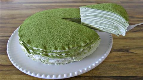 A matcha green tea crepe cake is placed on a white plate. A slice has been lifted from the cake, revealing its many layers filled with cream. The top of the cake is dusted with green matcha powder. The cake sits on a wooden table.