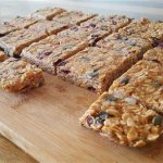 A wooden chopping board displays an array of rectangular homemade granola bars. The bars are filled with oats, nuts, seeds, and dried fruits, creating a colorful texture. The table is made of wood and adds a rustic feel to the presentation.