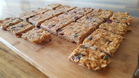 A wooden chopping board displays an array of rectangular homemade granola bars. The bars are filled with oats, nuts, seeds, and dried fruits, creating a colorful texture. The table is made of wood and adds a rustic feel to the presentation.